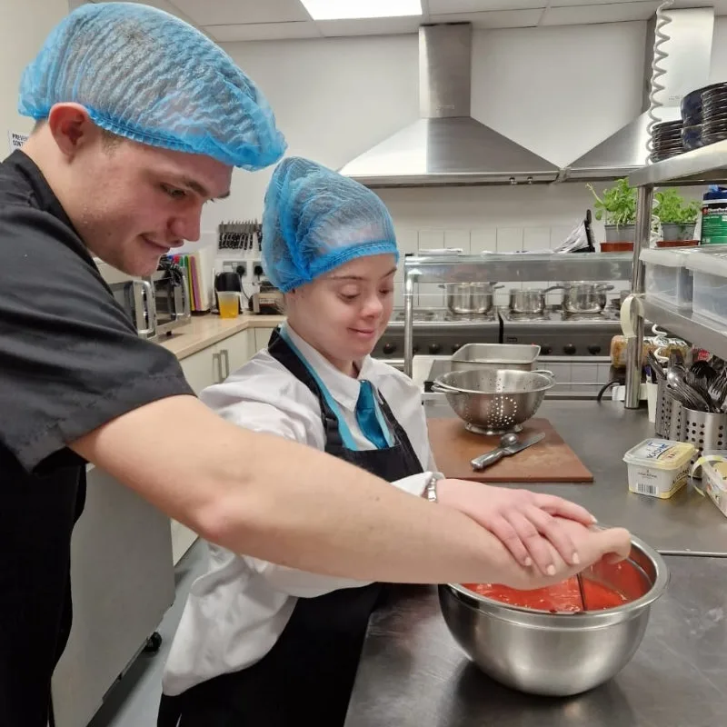 Two young people in hair nets and aprons mixing food in a steel bowl in a commercial kitchen