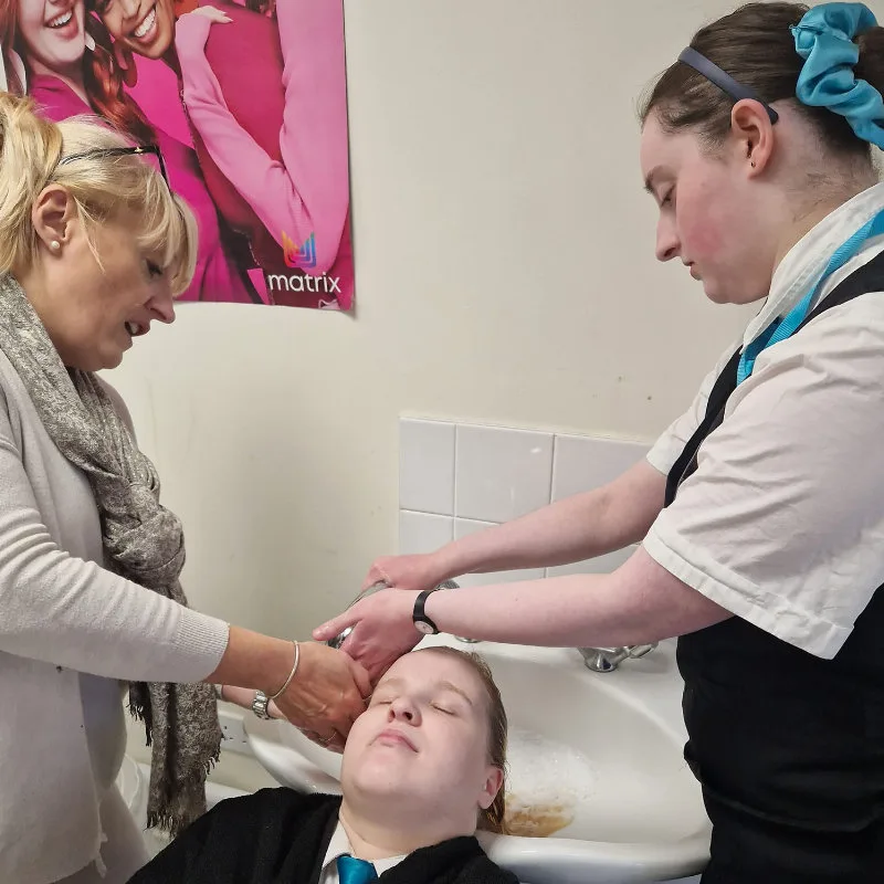 A trainee in a salon tunic washing a client's hair at a basin while a tutor watches