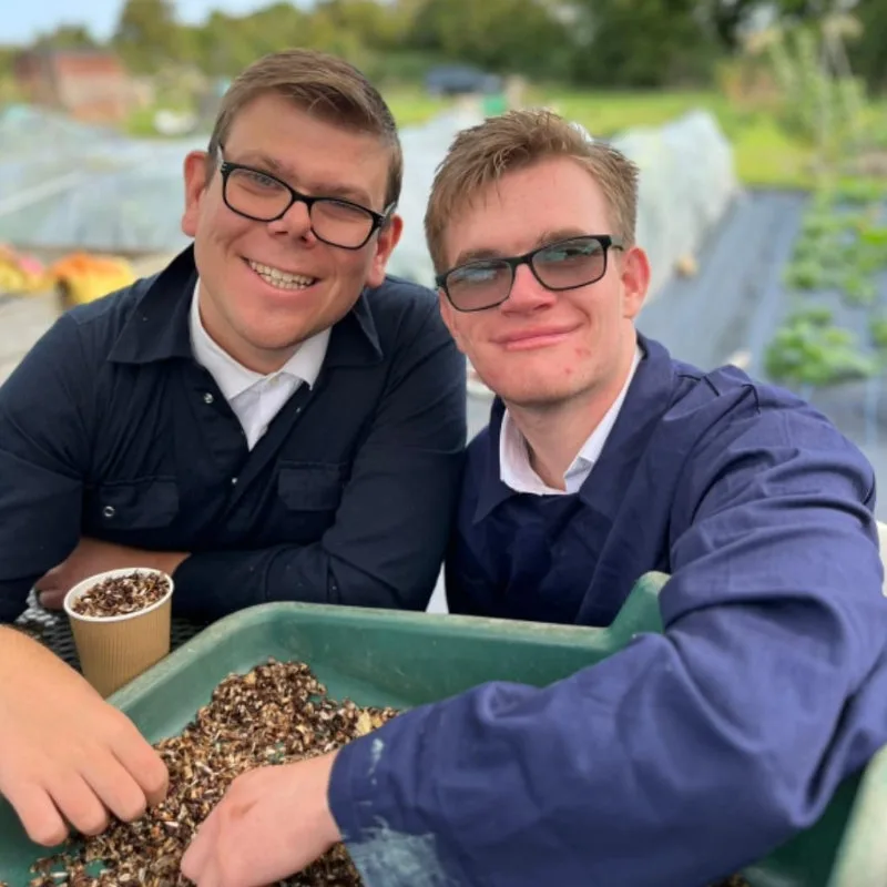 Two smiling young men in glasses leaning over a tray of seeds outdoors