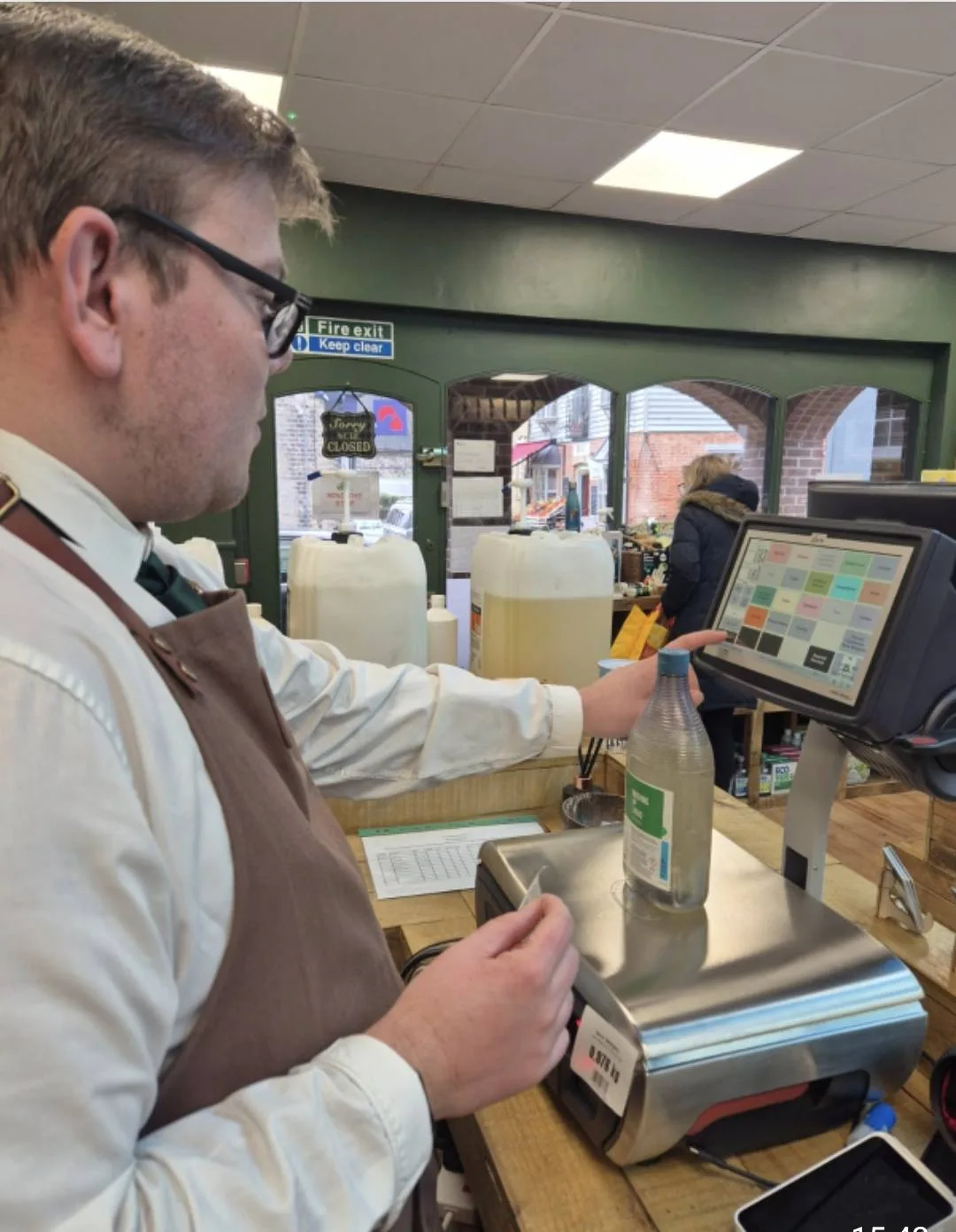 A man in an apron weighing a bottle of liquid on scales beside a till in a refill shop