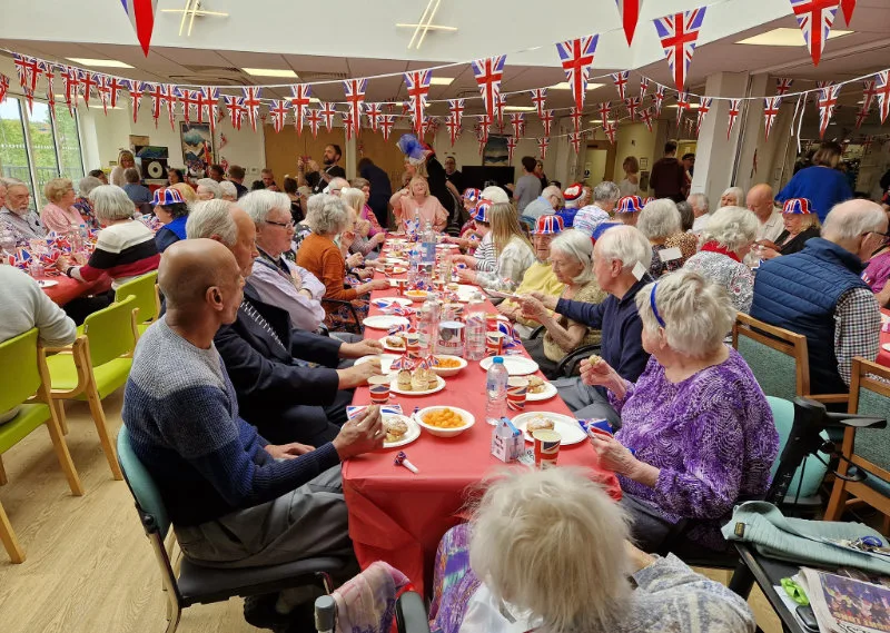 A long dining table of older guests under Union flag bunting at a community lunch