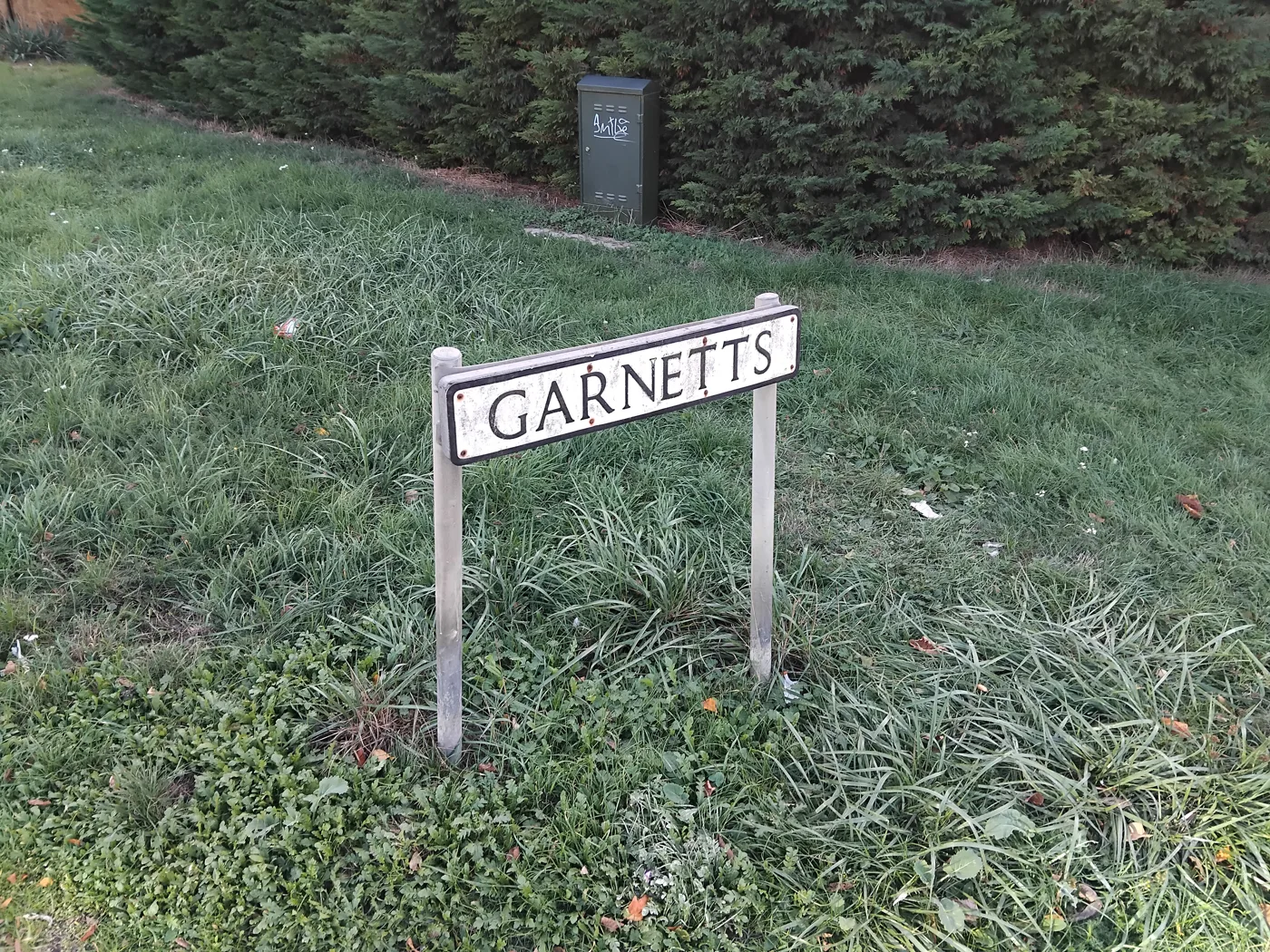 The Garnetts street name sign on a grass verge in Takeley, with a hedge and a green utility cabinet behind it